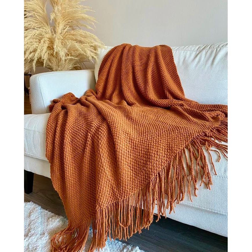 A rust-orange textured knit throw blanket with fringe, draped over a white sofa in a living room setting.