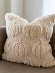 A square cream throw pillow with a textured, fuzzy knit design featuring raised pumpkins and leaves, resting on a brown sofa.