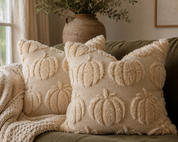 Two cream-colored textured throw pillows with raised pumpkin motifs, styled on an olive green couch with a matching knit throw blanket and a rustic vase in the background.