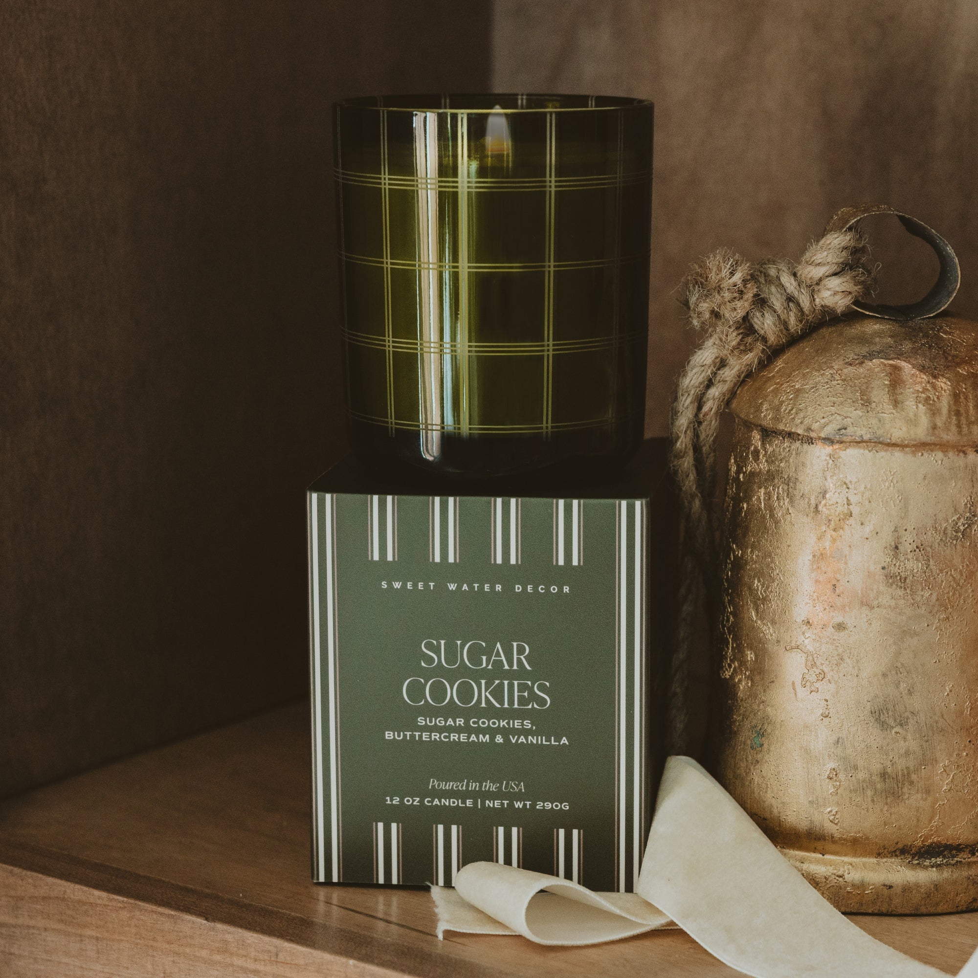 A candle product shown with its dark green glass jar and matching rectangular box on a wooden surface. The box reads 'SUGAR COOKIES' and 'Sweet Water Decor.