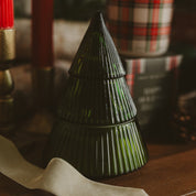 A dark green ribbed glass Christmas tree-shaped candle holder or decorative container, shown on a wooden surface with blurred holiday decor in the background.