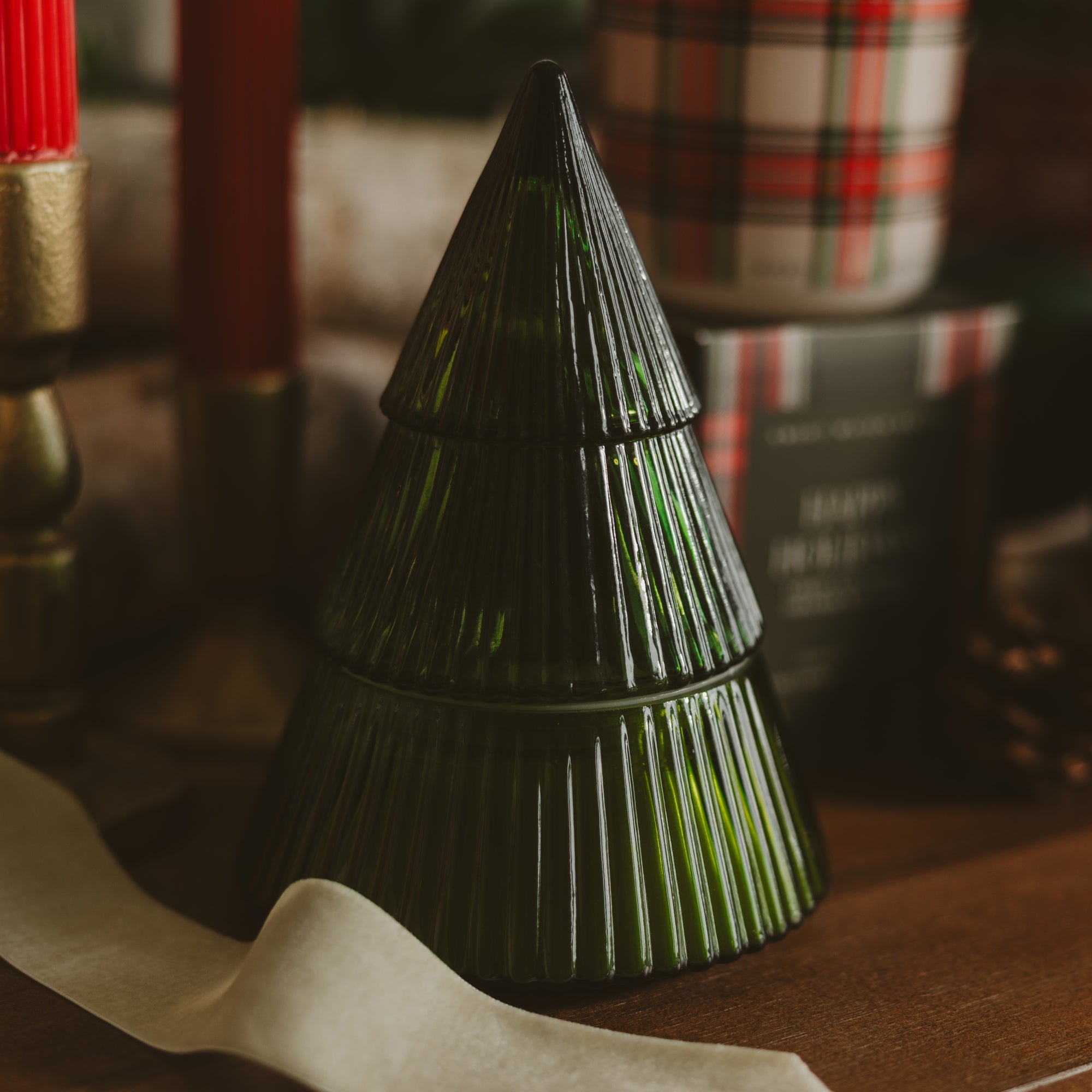 A dark green ribbed glass Christmas tree-shaped candle holder or decorative container, shown on a wooden surface with blurred holiday decor in the background.