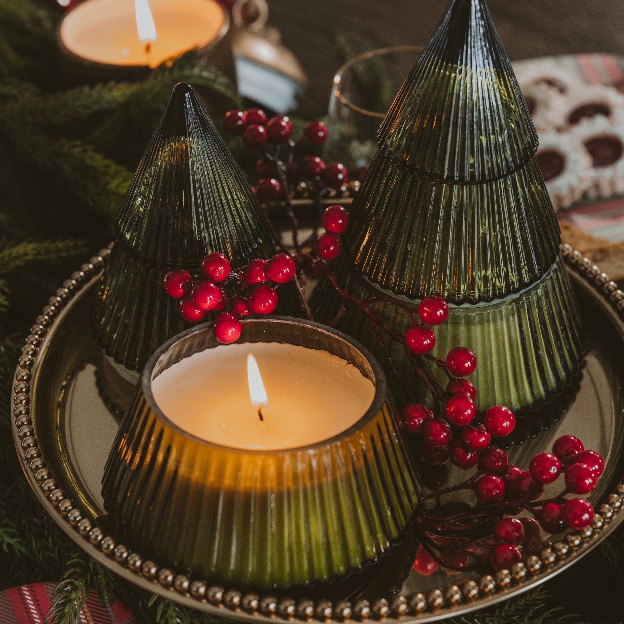 Three ribbed glass holiday tree candles in dark green shades, with two lit candles on a decorative silver tray surrounded by red berries and evergreen branches.
