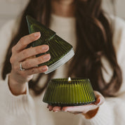 A person in a cream knit sweater holds an open ribbed dark green glass candle holder, showing the lit candle inside.
