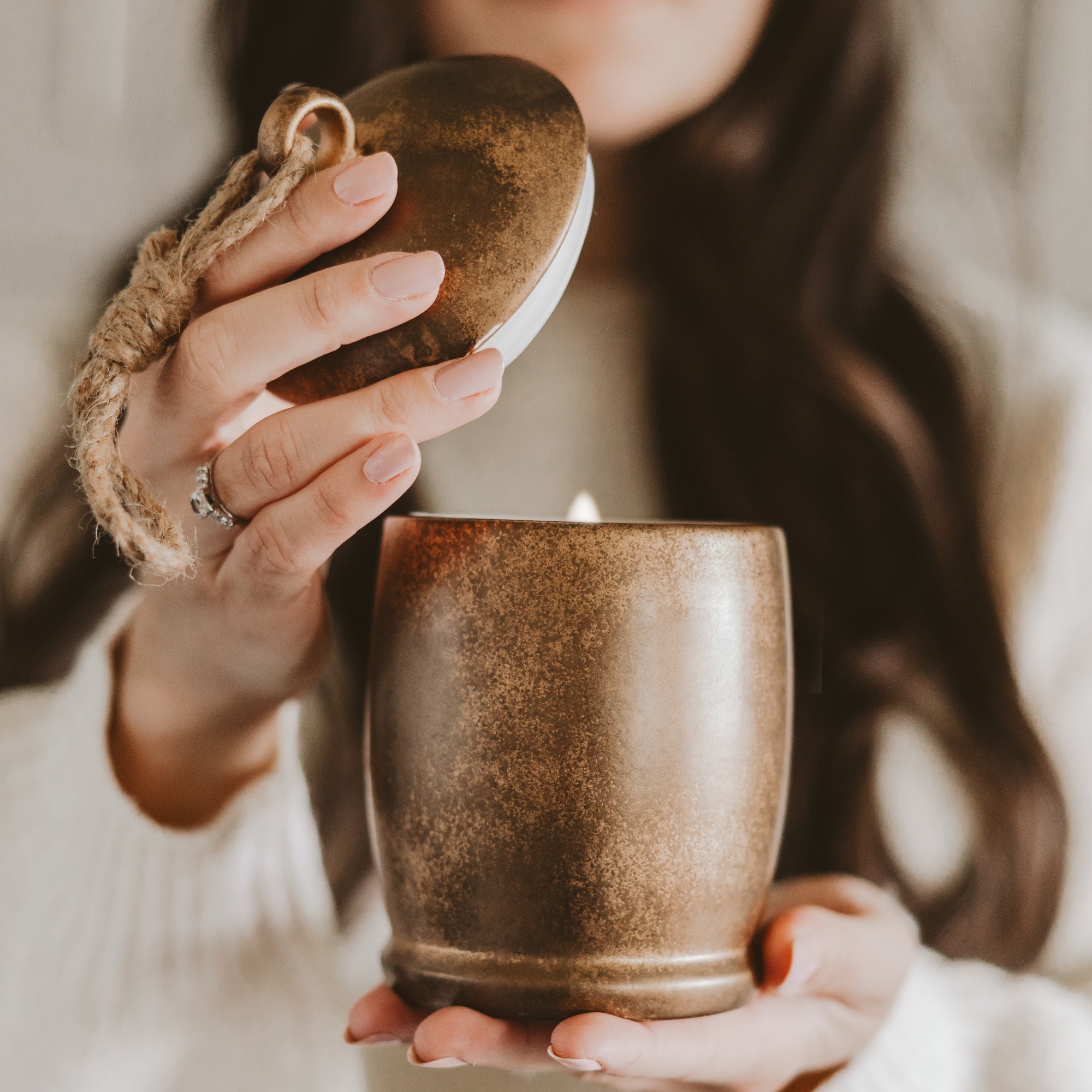 A person holds a lit candle in a rustic brown ceramic holder, lifting its matching brown lid with a jute loop. The person is wearing a white knit sweater, and the background is softly blurred.