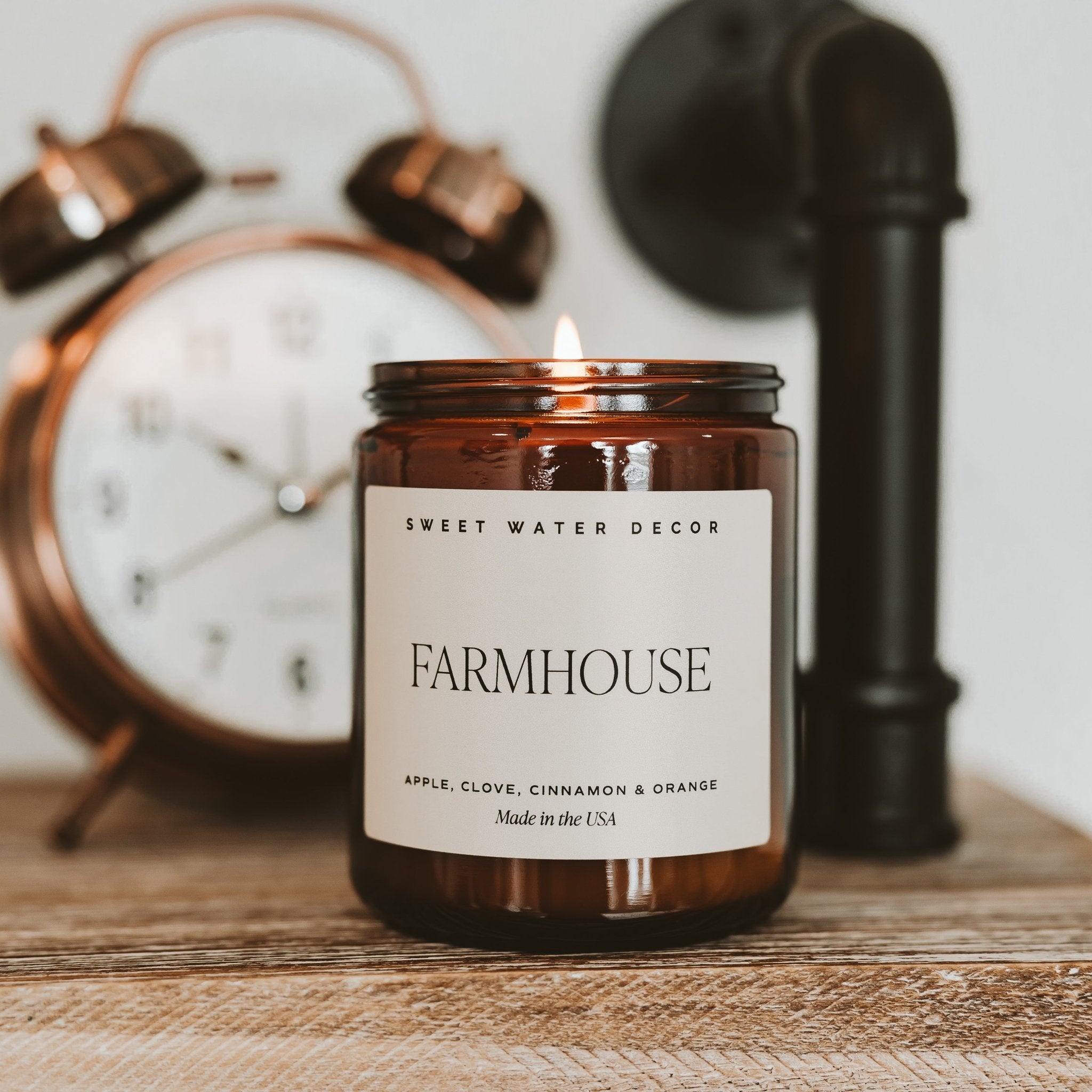Candle labeled 'Farmhouse' by Sweet Water Decor with an alarm clock and pipe in the background.