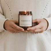 Person holding a 'Farmhouse' candle in a textured sweater