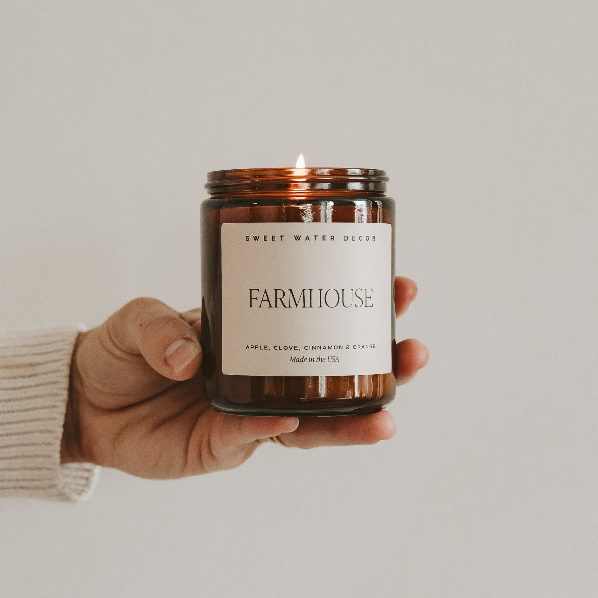 Hand holding a 'Farmhouse' candle with a visible label against a neutral background
