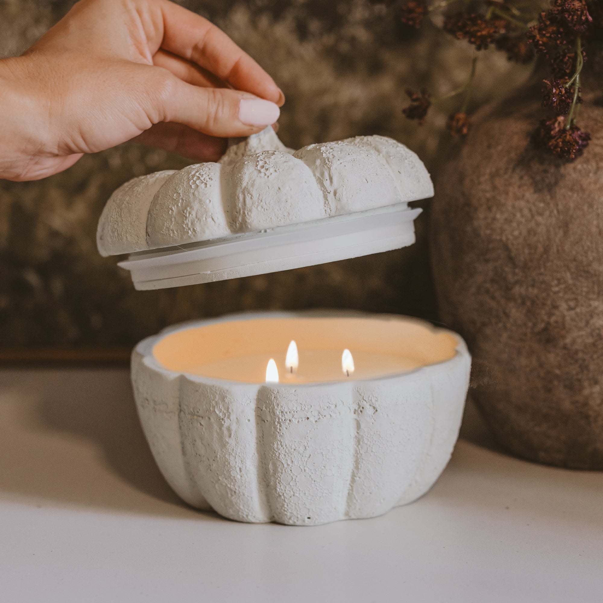A hand lifts the lid of a white textured pumpkin-shaped candle holder containing a lit candle, with a rustic brown vase in the background.