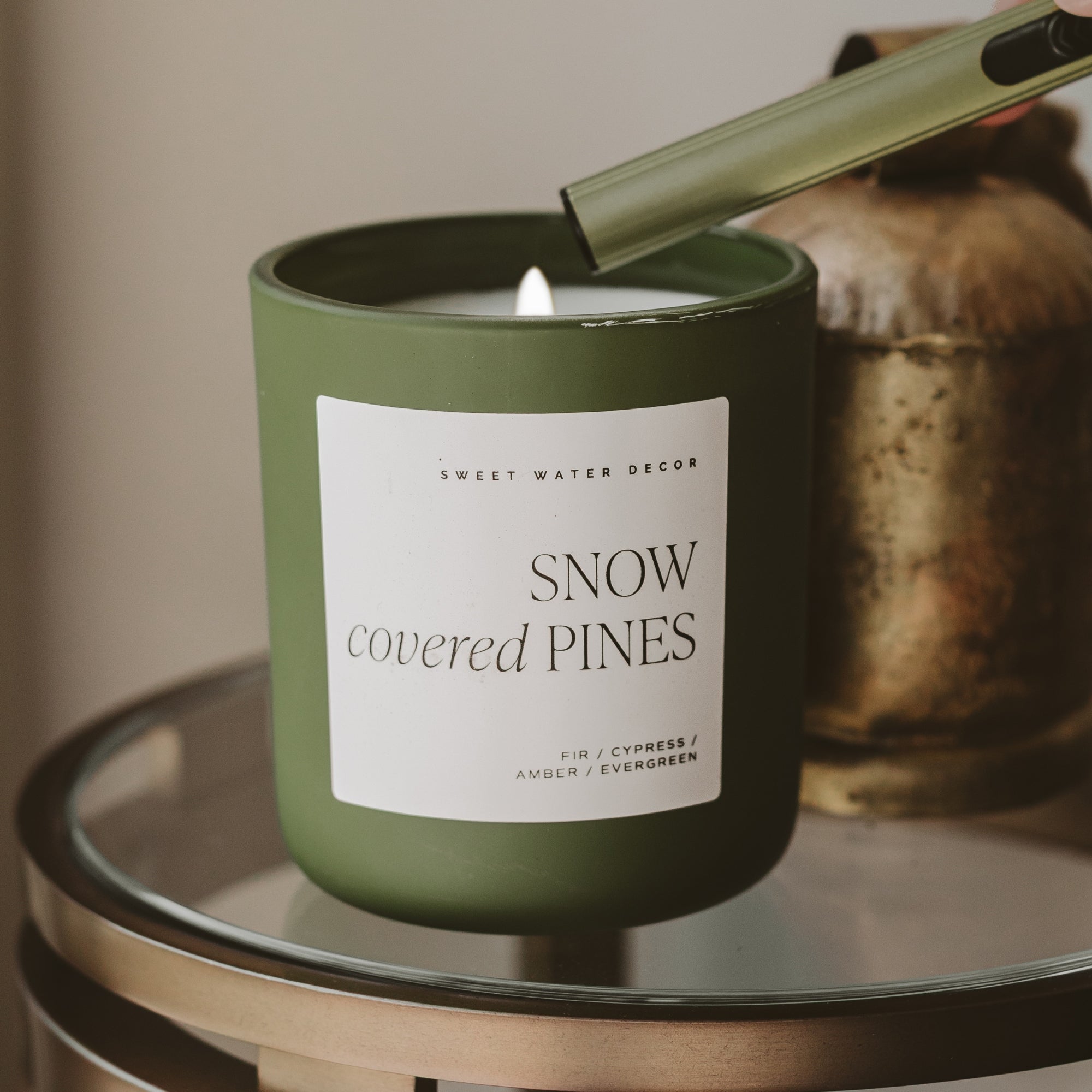 An olive green candle jar labeled 'SNOW covered PINES' by Sweet Water Decor, lit with a visible flame and topped with a matching green wick holder, set on a glass table with a blurred rustic vessel in the background.