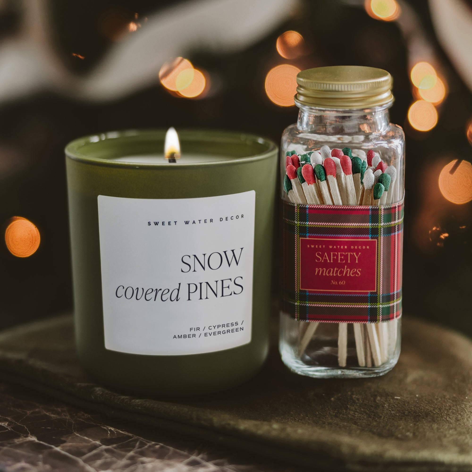 A lit olive green candle labeled 'SNOW covered PINES' by Sweet Water Design sits beside a glass jar of 'SAFETY matches' with red and green striped matches, set on a dark surface with warm bokeh lights in the background.