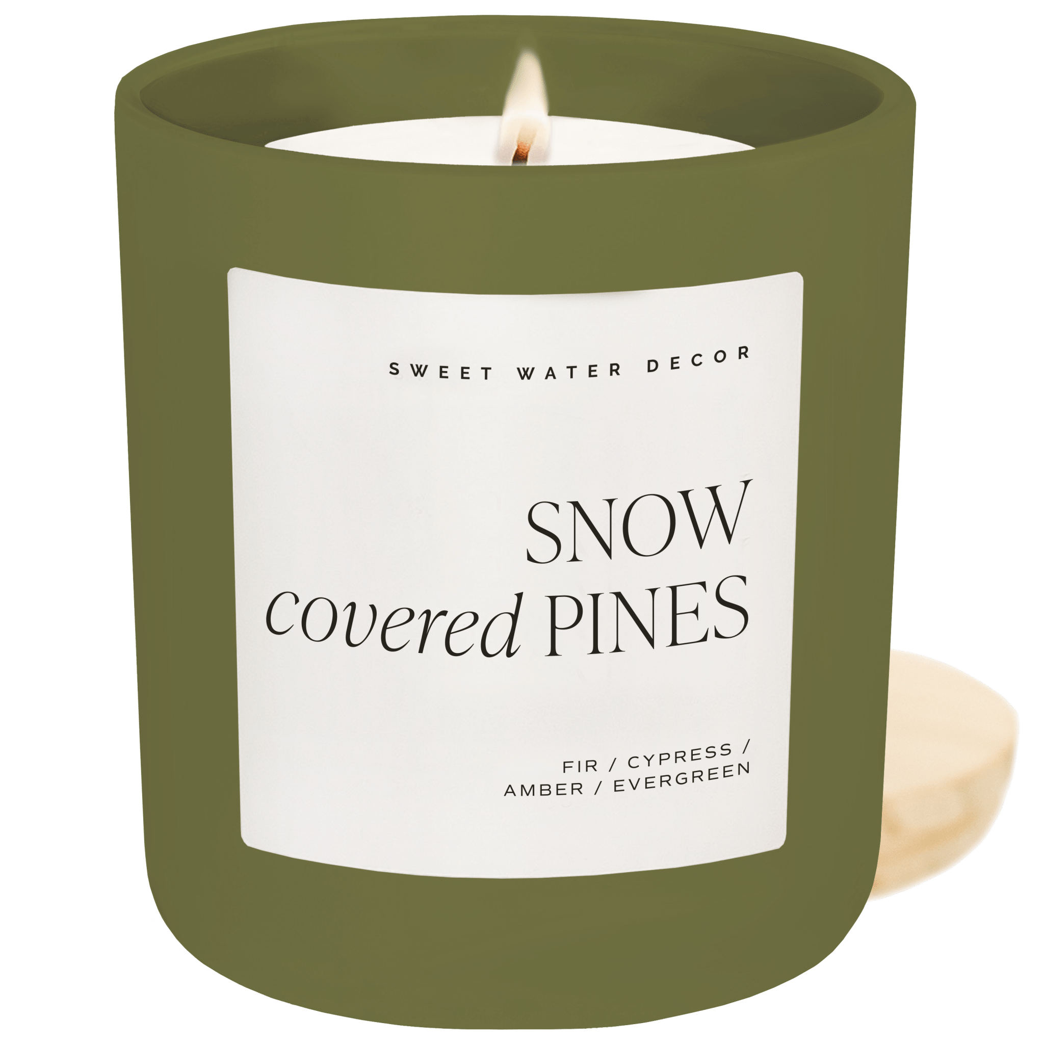 A lit candle in an olive green glass jar with a white label reading 'SNOW covered PINES' and 'SWEET WATER DECOR' at the top, shown against a matching olive green background.