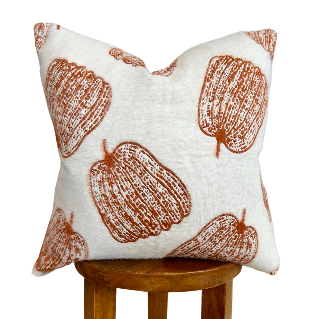 A square throw pillow with a white linen-like background and a rust-orange pumpkin print, displayed on a wooden stool against a white background.
