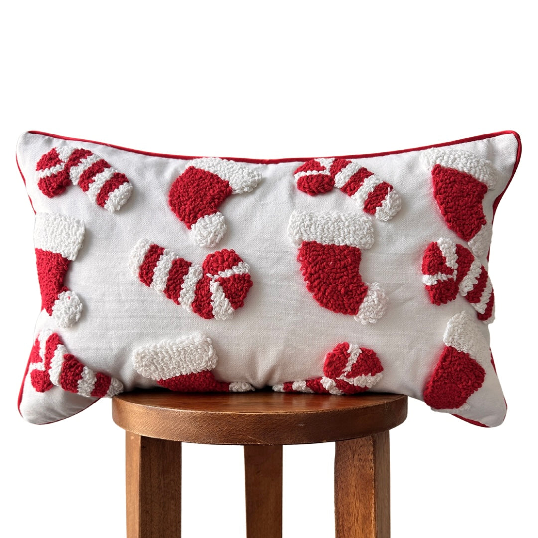 A rectangular white throw pillow with red and white textured holiday motifs, including candy canes and stockings, displayed on a wooden stool against a white background.