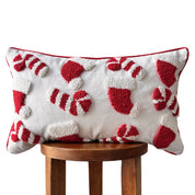 A rectangular white throw pillow with red and white textured holiday motifs, including candy canes and stockings, displayed on a wooden stool against a white background.