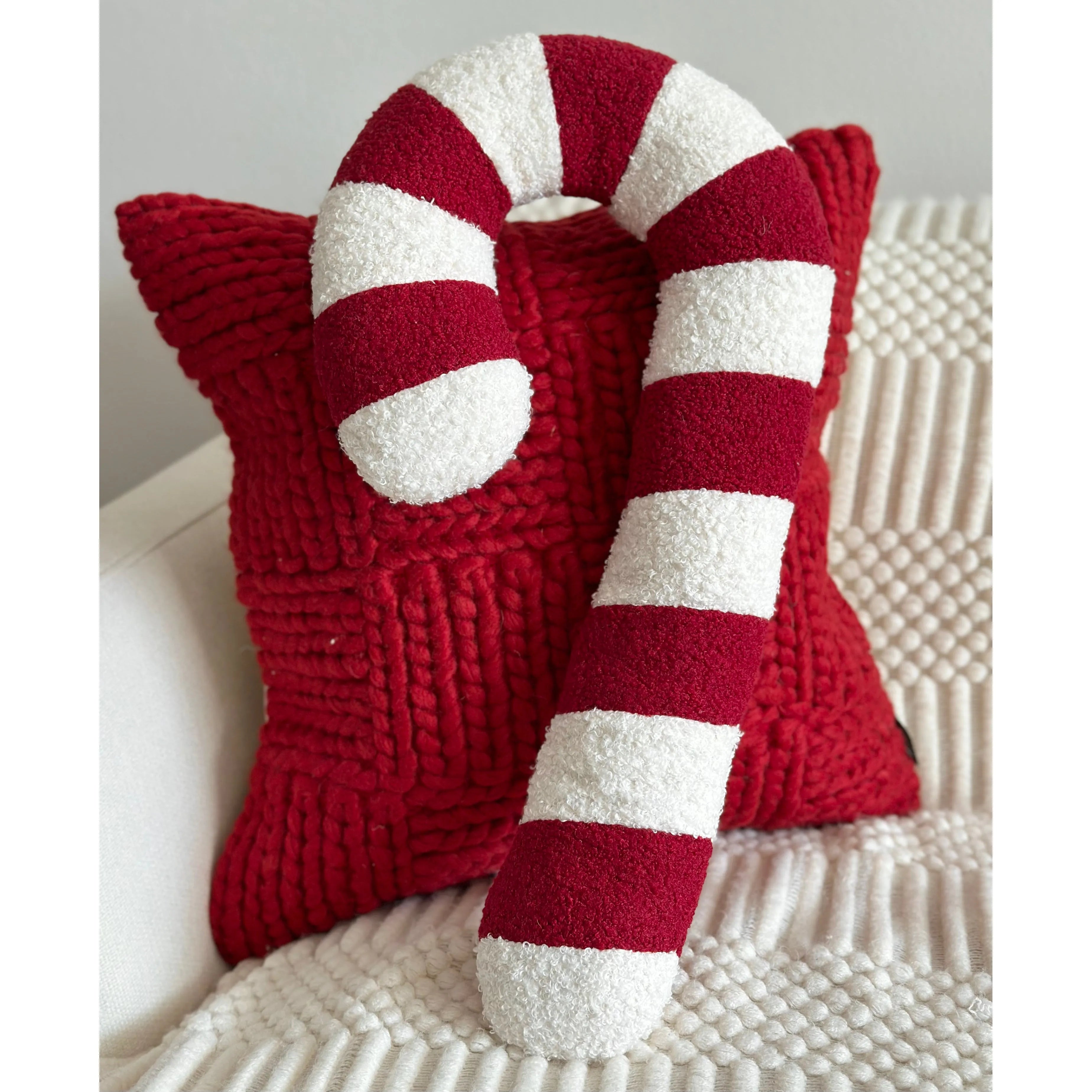 A red and white striped candy cane plush pillow resting on top of a textured red star-shaped throw pillow, set on a cream knit blanket.