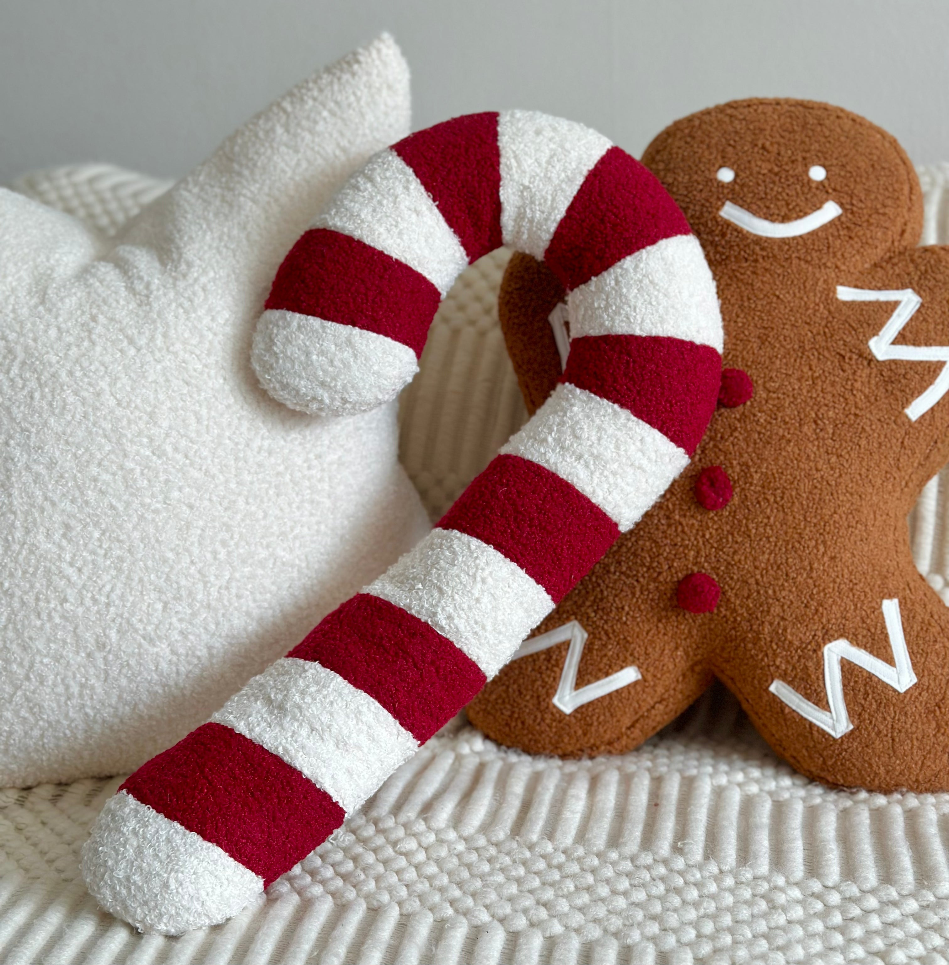 A plush holiday-themed pillow set featuring a large red and white striped candy cane and a brown gingerbread man with white icing details, resting on a white textured blanket.