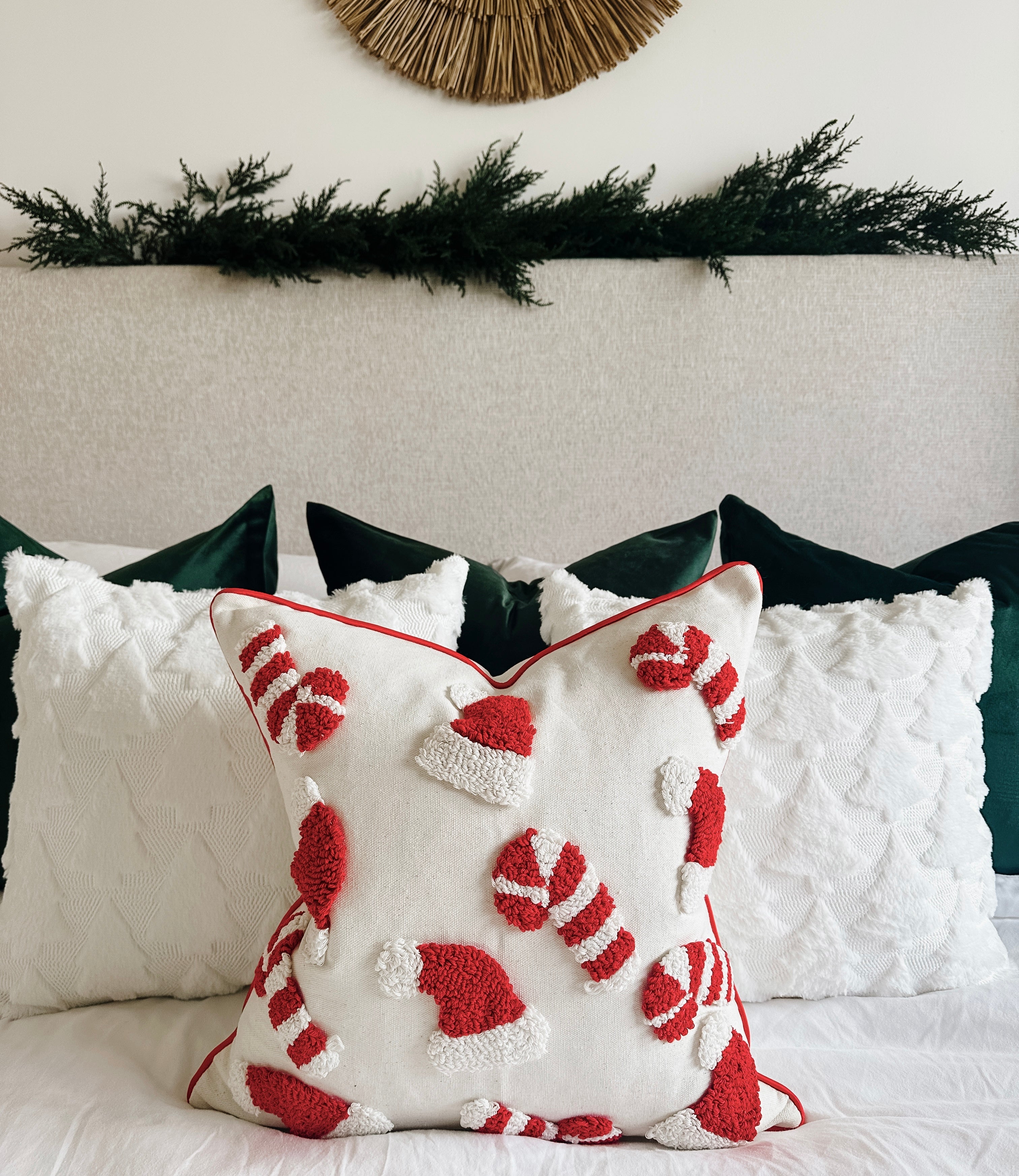 A festive Christmas throw pillow with red and white candy cane and Santa hat motifs, styled on a bed with white and dark green pillows against a neutral headboard.