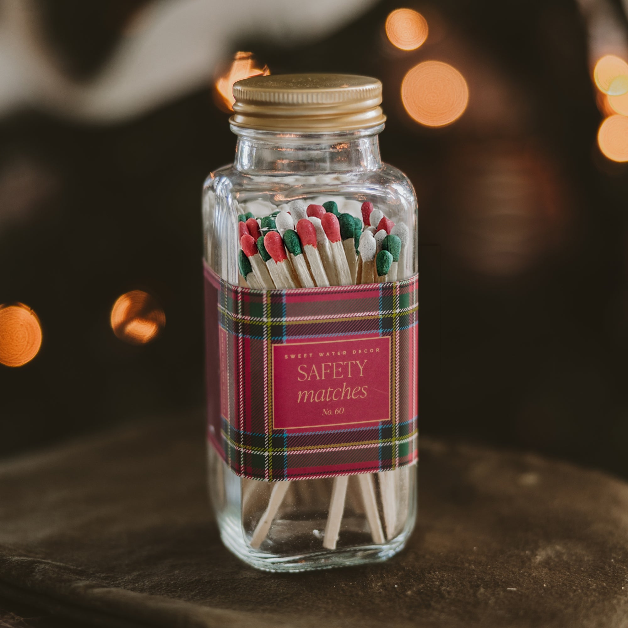 A clear glass bottle filled with wooden safety matches, sealed with a gold metal cap and wrapped in a red plaid label reading 'SAFETY matches' and 'Sweet Water Supply No. 82.