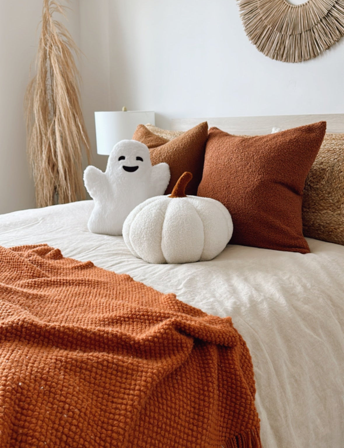 Cozy bedroom with a bed featuring orange pillows and a white plush ghost, with a decorative wall hanging.