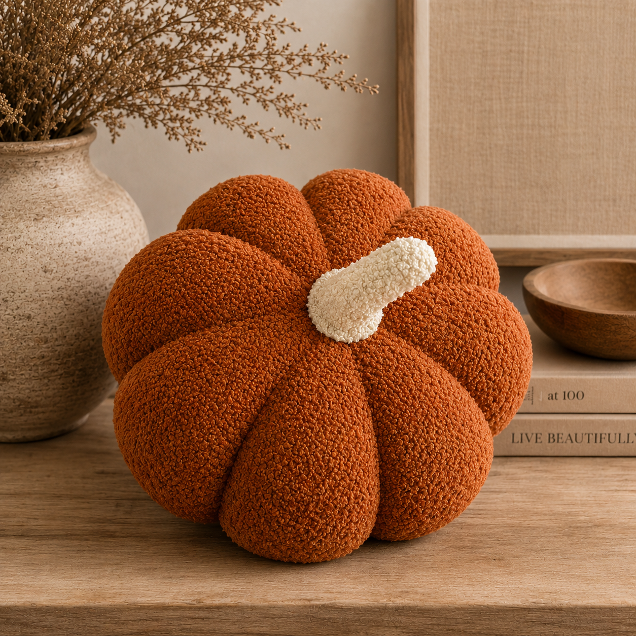 A textured, ribbed orange pumpkin-shaped decorative pillow with a cream-colored stem, styled on a wooden table with a ceramic vase, dried branches, and a book in the background.