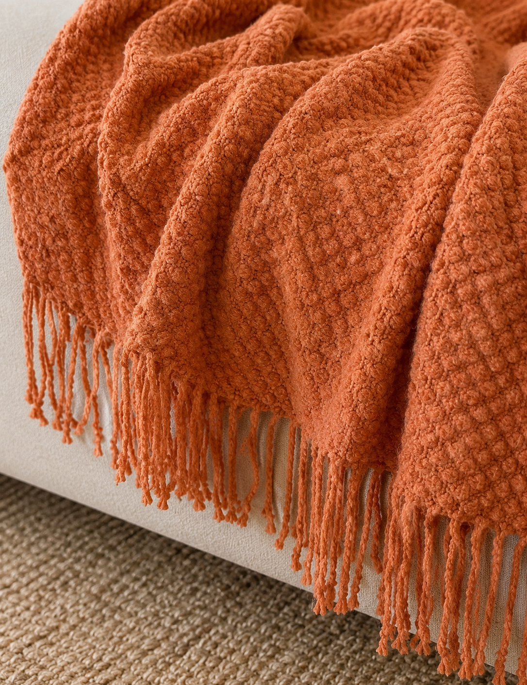 A close-up of a textured terracotta orange throw blanket with fringe, draped over a light beige upholstered armchair on a woven rug.