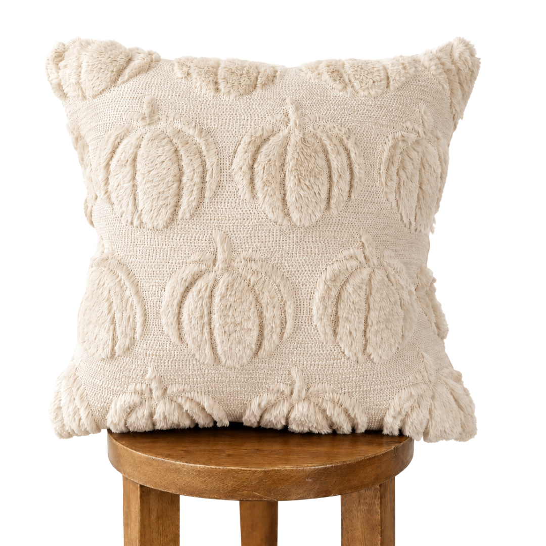 A square cream throw pillow with a raised, textured pumpkin pattern, resting on a wooden stool against a solid white background.