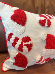 A white throw pillow with red trim and raised red-and-white holiday motifs, including Santa hats, candy canes, and mittens, shown resting on a brown leather couch.
