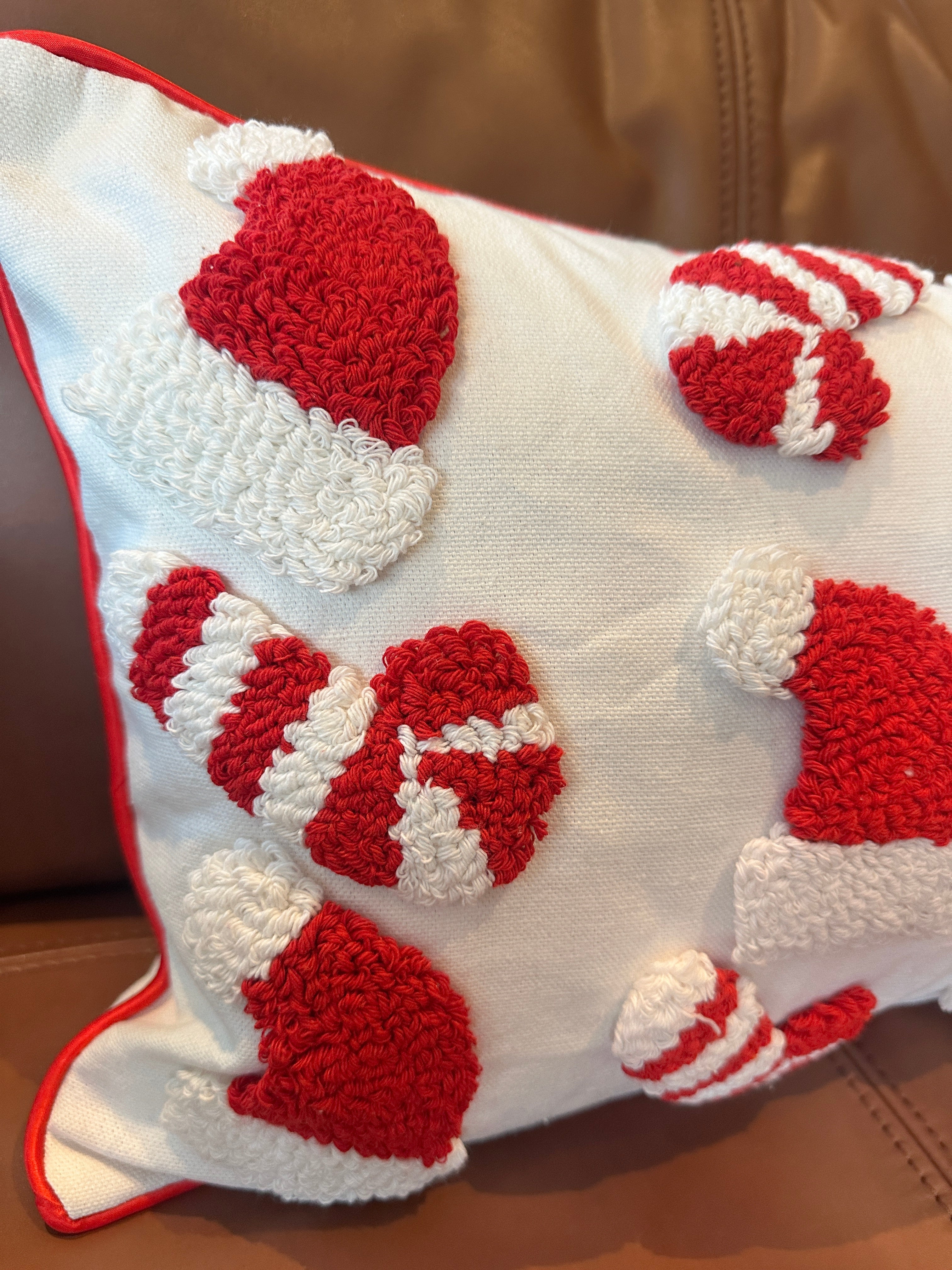 A white throw pillow with red trim and raised red-and-white holiday motifs, including Santa hats, candy canes, and mittens, shown resting on a brown leather couch.