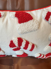 A close-up of a textured throw pillow featuring red and white Christmas-themed tufted motifs, including Santa hats and candy canes, with a red piped border, resting on a brown leather couch.
