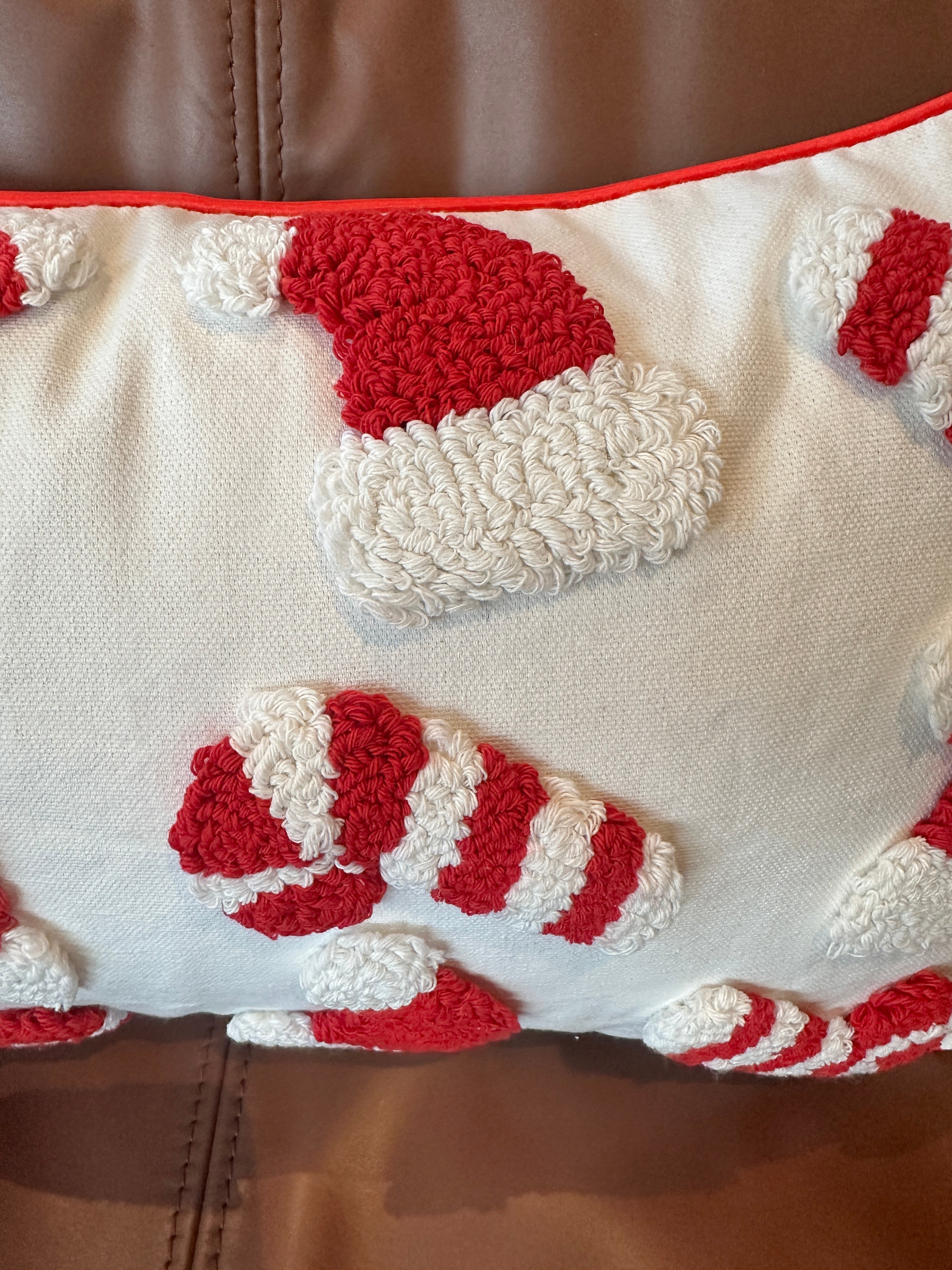A close-up of a textured throw pillow featuring red and white Christmas-themed tufted motifs, including Santa hats and candy canes, with a red piped border, resting on a brown leather couch.