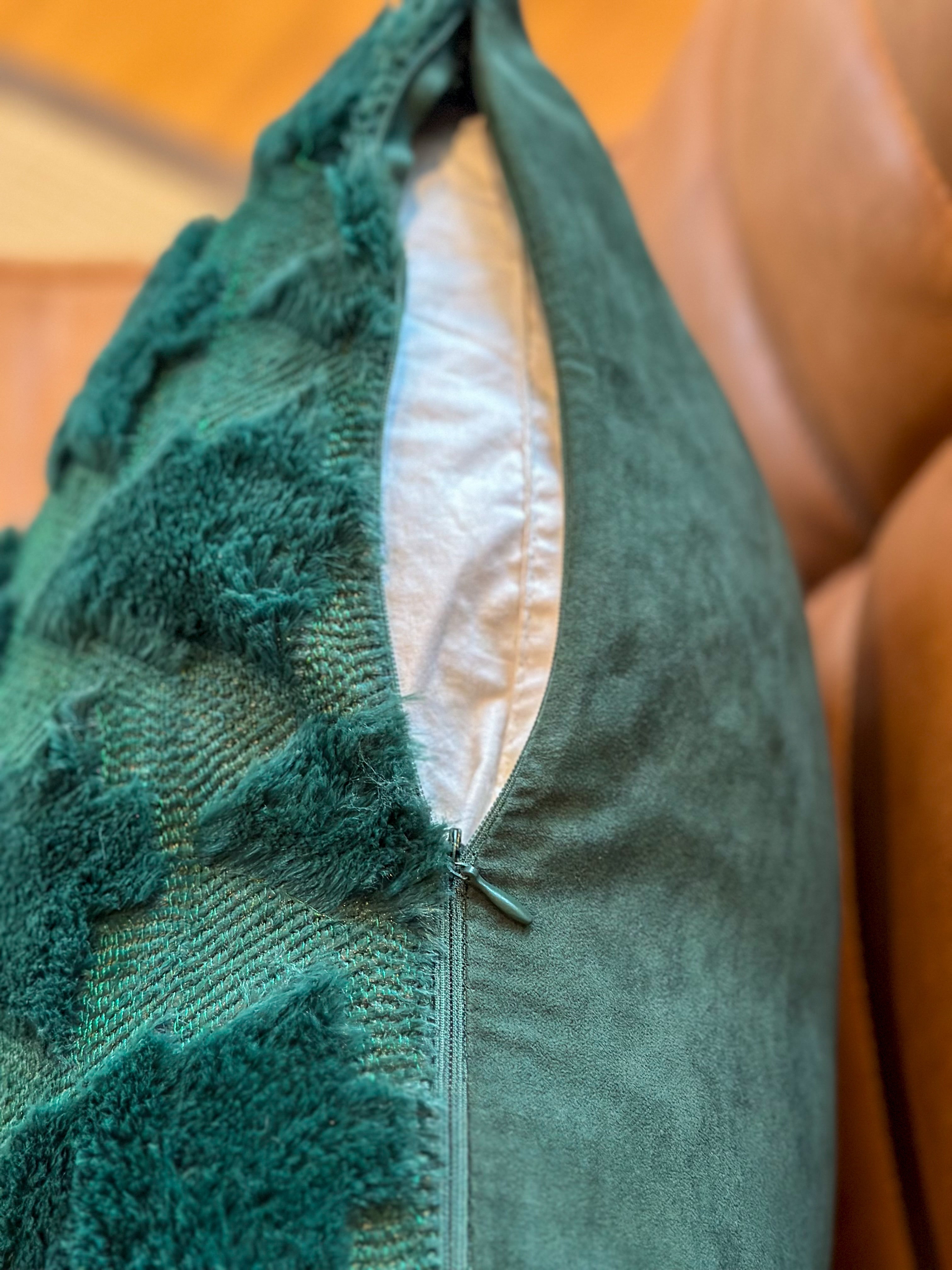 A close-up of a textured teal throw pillow with a zippered opening, showing a white inner lining, resting on a brown leather sofa.