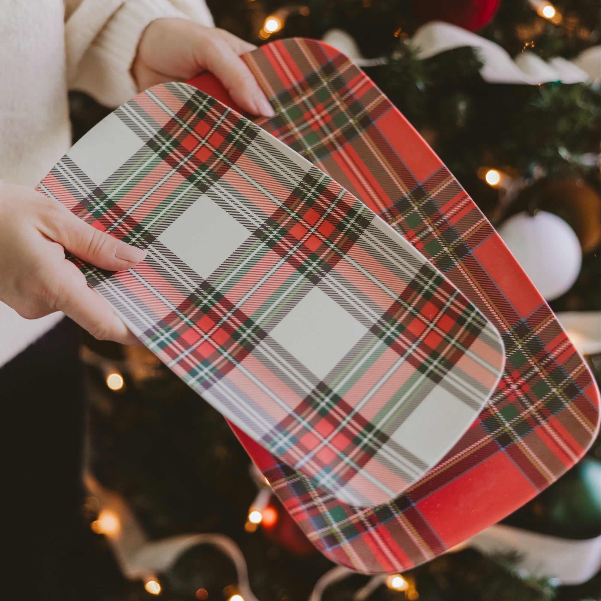 A person holds a rectangular platter with rounded corners featuring a red, green, black, and white tartan plaid pattern, with a second matching platter underneath. The background shows a softly blurred Christmas tree with warm lights and ornaments.