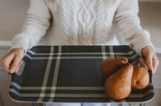 A person in a cream cable-knit sweater holds a rectangular plaid tray with dark gray and beige stripes. On the tray are three brown pears.