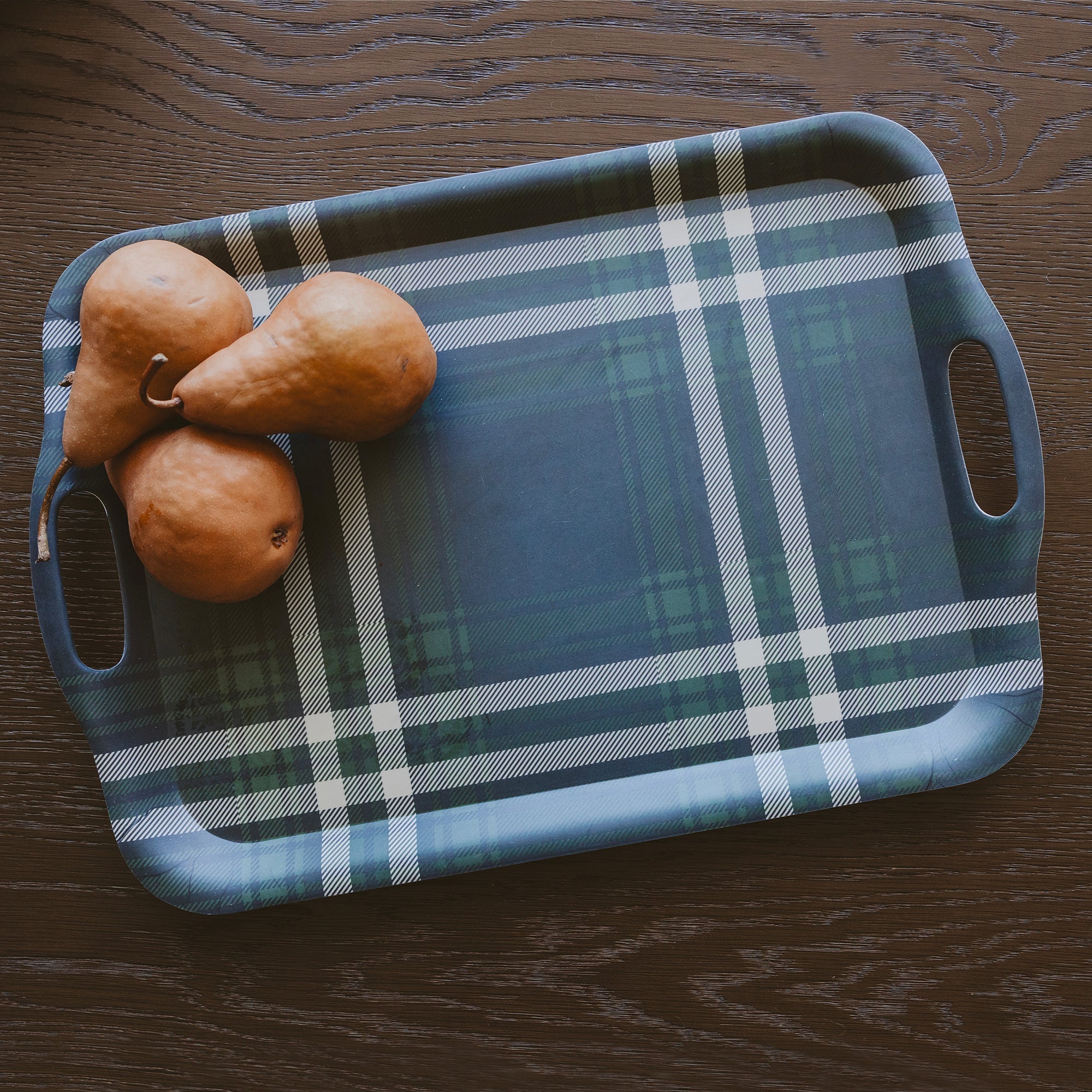 A rectangular plaid serving tray with cutout handles sits on a dark wood tabletop, with three brown pears placed on the left side of the tray.