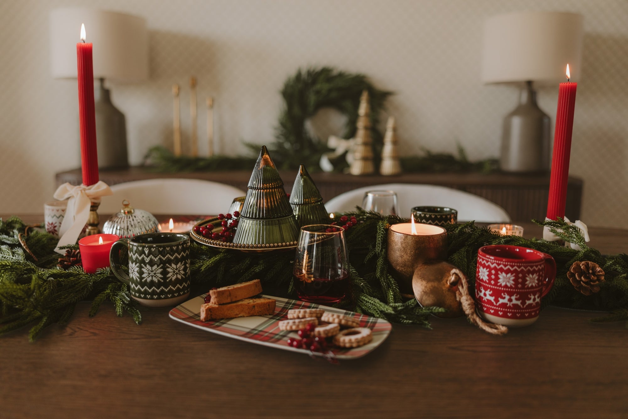 A cozy holiday table setting with red candles, green pine garlands, patterned ceramic mugs, small decorative Christmas trees, and a platter of cookies on a wooden table in a warmly lit dining room.