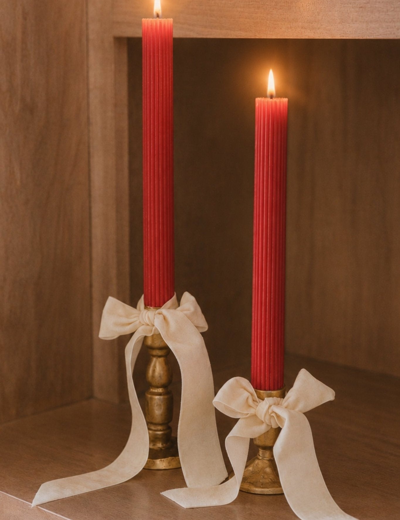 Two tall red taper candles in brass holders, tied with cream ribbon bows, displayed on a wooden shelf with both candles lit.