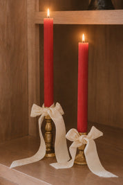 Two tall red taper candles in brass holders, tied with cream ribbon bows, displayed on a wooden shelf with both candles lit.