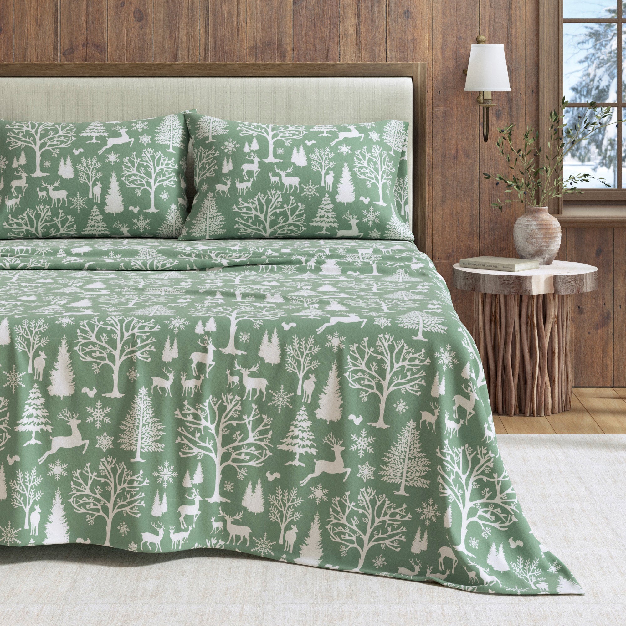 Green forest-themed bedspread on a bed with matching pillows in a room with wooden walls and a lamp.