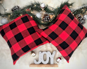 Red and black checkered pillow with Christmas decorations including pine cones and 'JOY' sign.