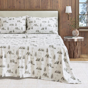 Bed with bear and tree patterned bedding in a room with wooden walls and a lamp.