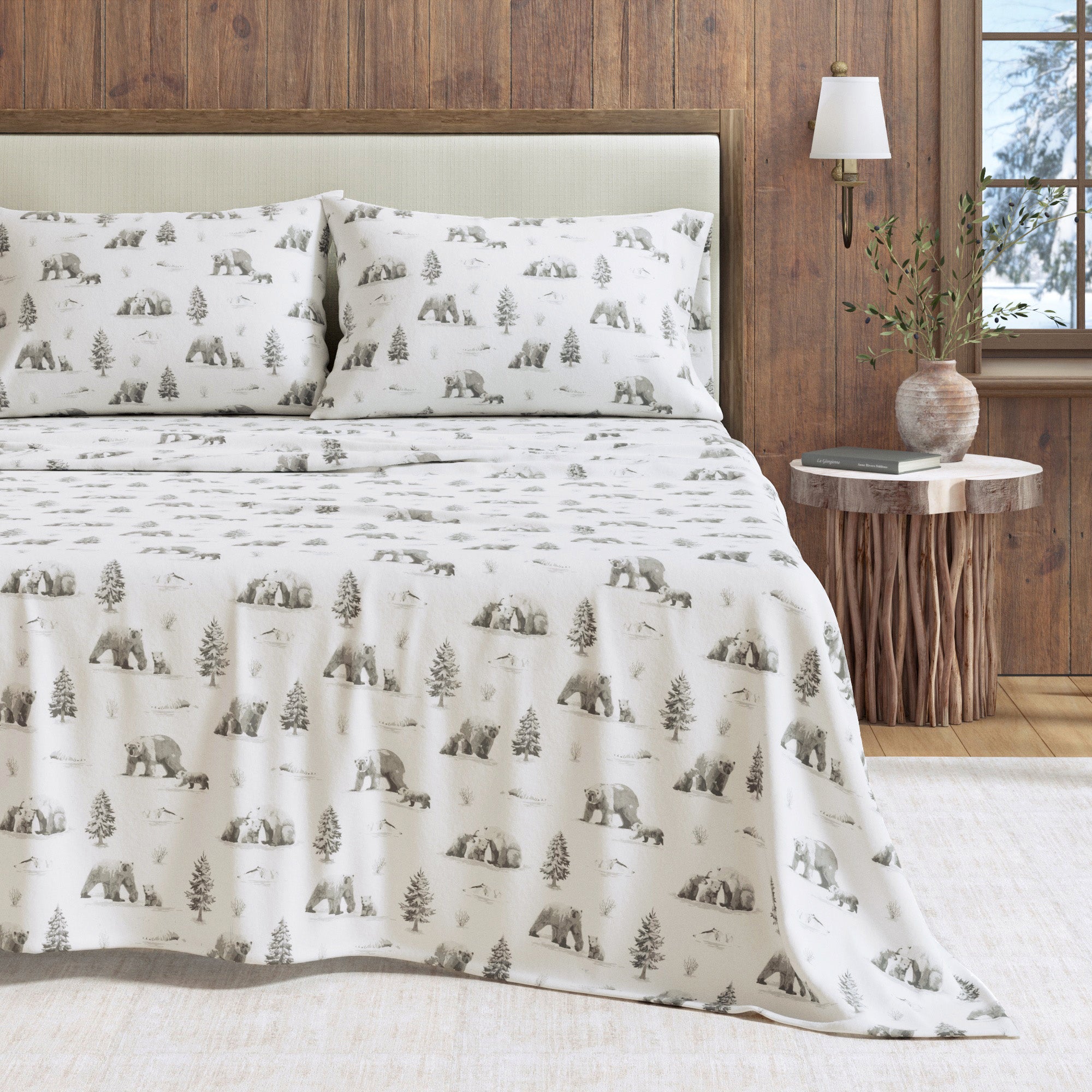 Bed with bear and tree patterned bedding in a room with wooden walls and a lamp.
