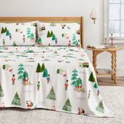 Christmas-themed bedspread with trees and people on a bed in a room with a lamp and table.
